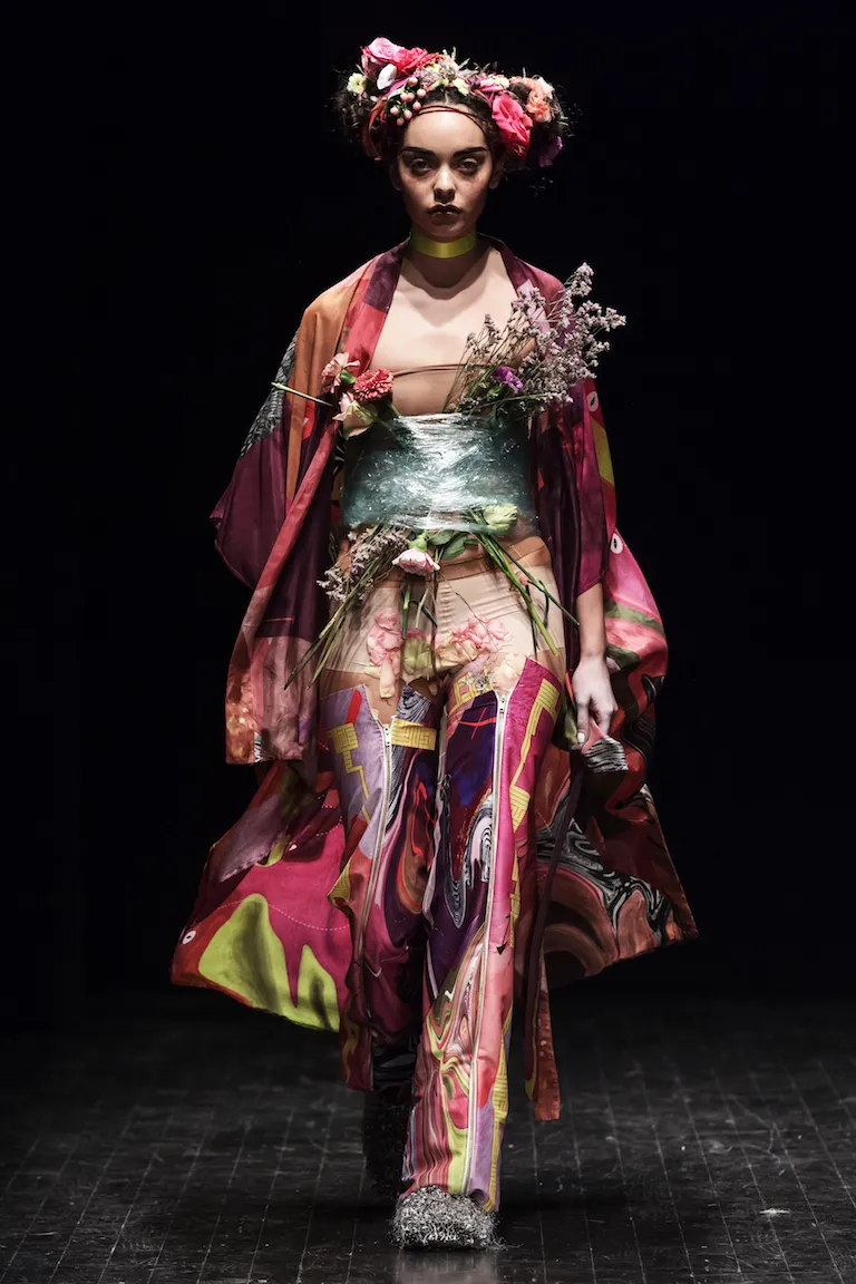 A floral crown and a transparent corset filled with flowers, worn over a coat and trousers in red, purple and lime green.