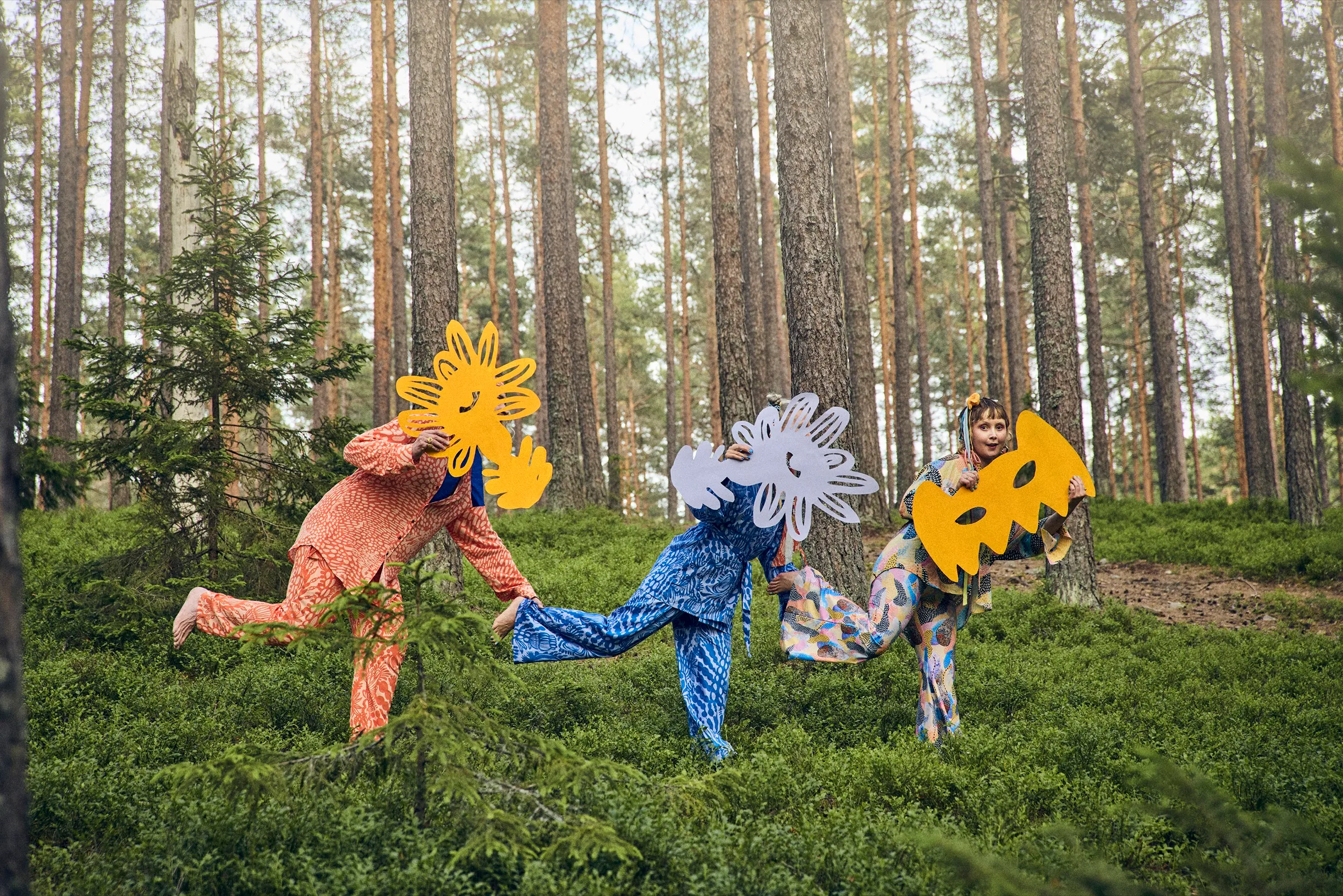 Tre personer i mönstrade set i rörelse över blåbärsris, var och en med en stor utklippt blomma framför ansiktet, Studio L'Narte