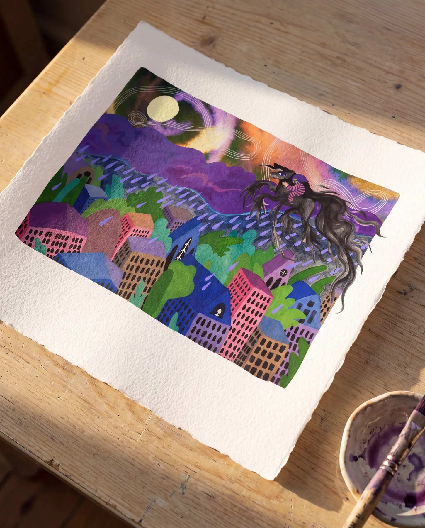 Originalet till uppslaget — gouache på akvarellpapper med riven kant — liggande på ett solbelyst arbetsbord i trä med en färgskål och pensel i lila bredvid: barnet på den svarta hästen flyger genom lila regn över staden med prickiga hus, under lila moln och fullmåne — verk av Amandah L'Narte