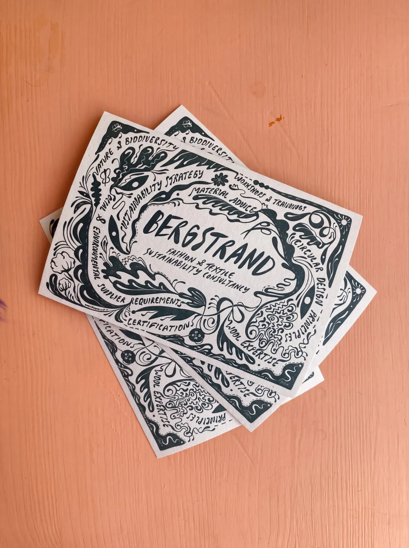 Fyra tryckta kort utlagda i solfjäder på en orangemålad träyta — mörkgrön illustration på benvitt papper där Bergstrand fashion & textile sustainability consultancy står handtextat i mitten, omgivet av en krans av blad, slingor och ögonformer med konsultbyråns tjänster textade runtom — uppdrag av Amandah L'Narte för Bergstrand Consultancy