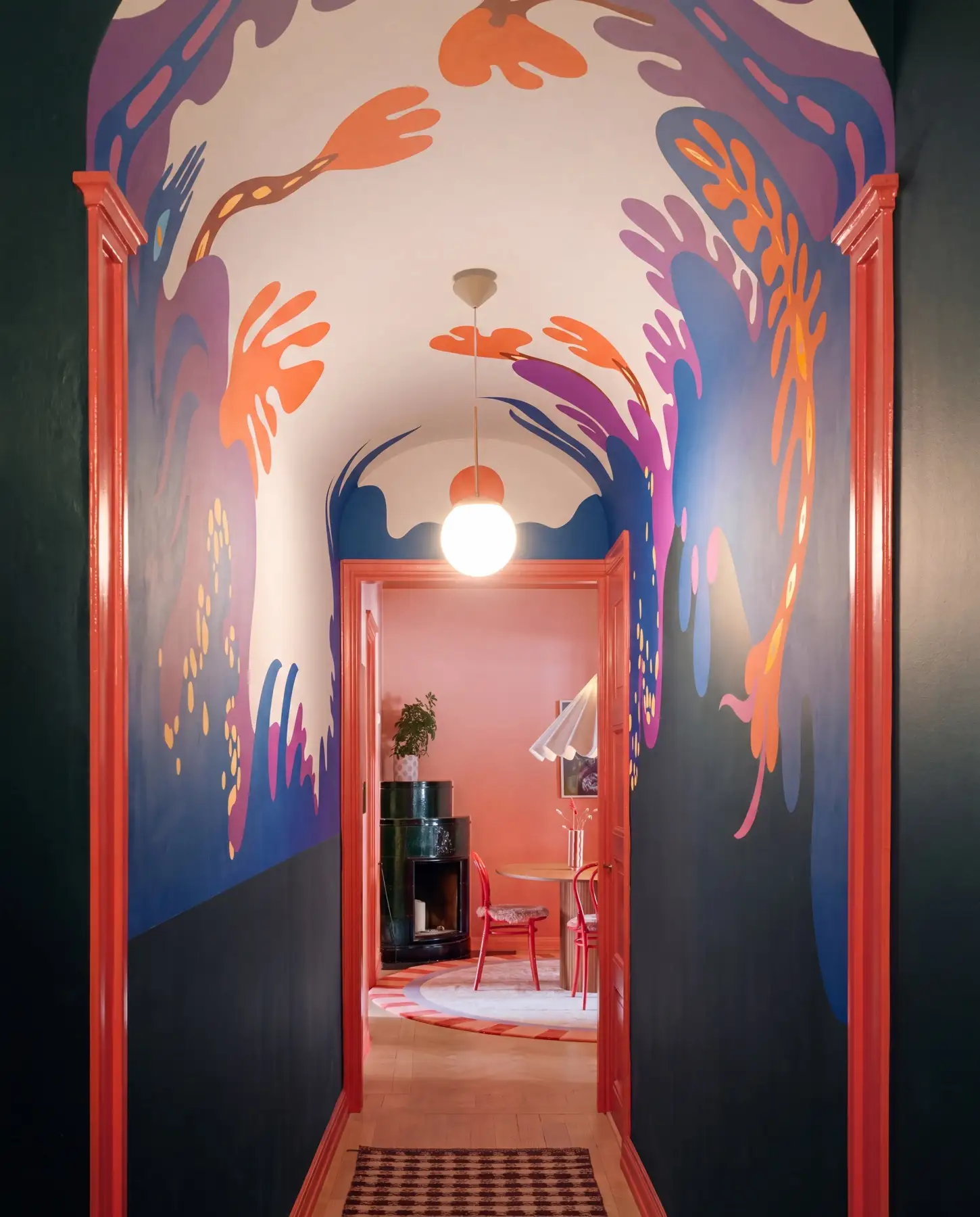 Valvslagen hall med muralmålning över tak och övre väggar: flödande växt- och korallformer i orange, lila, magenta och blått mot benvit botten, över mörkgröna nedre väggfält och korallröda dörrfoder — i fonden ett korallrosa rum med svart rund kamin, röda stolar och klotlampa — muralmålning av Amandah L'Narte för Sara Garanty