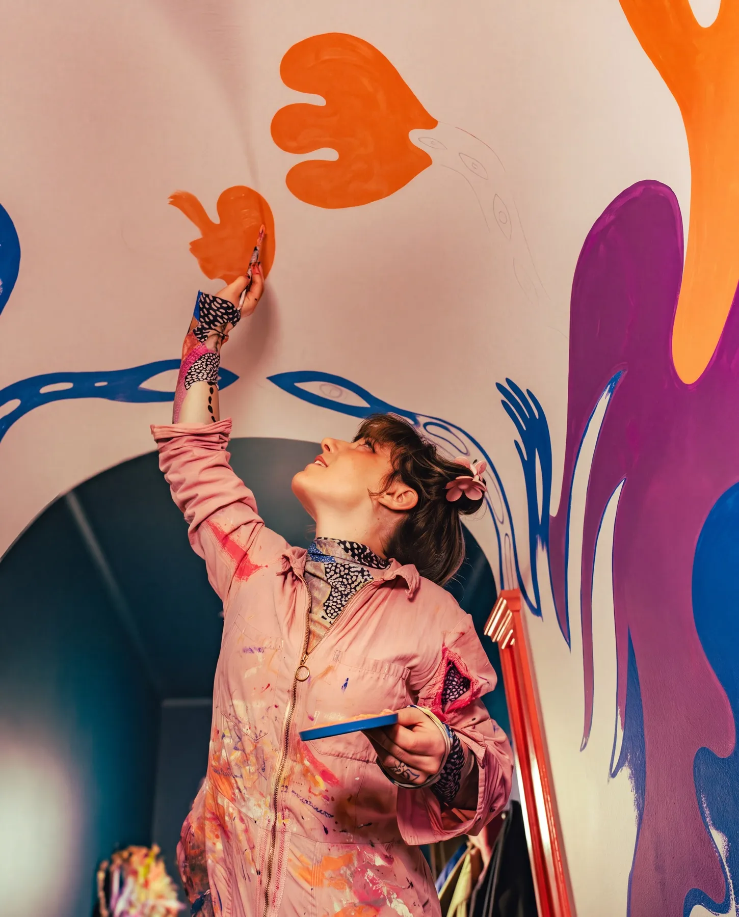 Amandah L'Narte i rosa färgstänkt overall målar en orange bladform i hallens valvtak med skisslinjer i blyerts ännu synliga bredvid — processbild från muralmålningen hos Sara Garanty