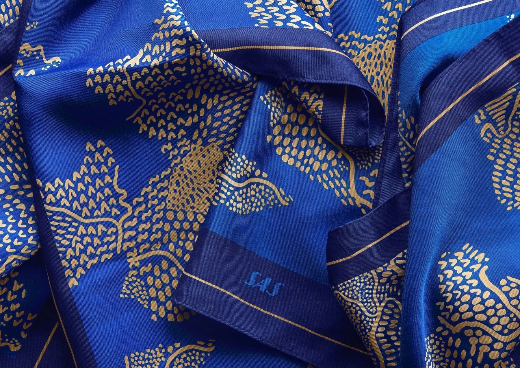 Klarblå scarf med mönster i guld — organiska träd- och landskapsformer tecknade i punkter och streck, mörkblå bård med SAS logotyp — uppdrag av Amandah L'Narte för SAS