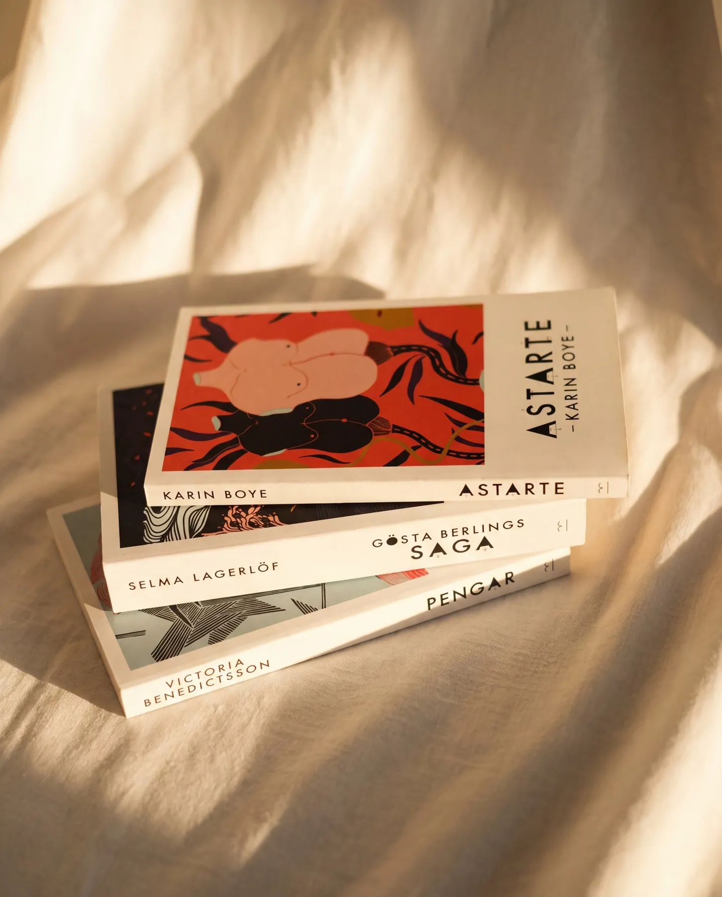 Seriens tre böcker i trave med ryggarna mot betraktaren på skrynklat linnetyg i varmt sidoljus: Karin Boyes Astarte överst, därunder Selma Lagerlöfs Gösta Berlings saga och Victoria Benedictssons Pengar — bokomslag av Amandah L'Narte för Trut Publishing