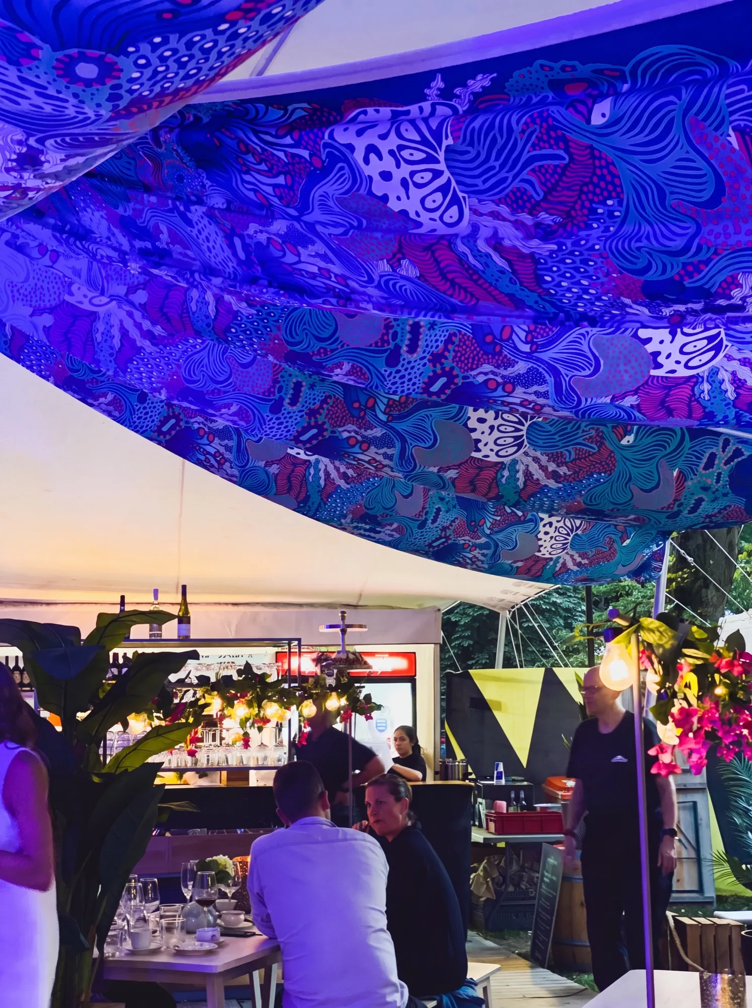 Coral Garden forms a fabric canopy above an outdoor bar at dusk, with cobalt-blue and coral-pink lengths stretched over guests seated beneath strings of lights.