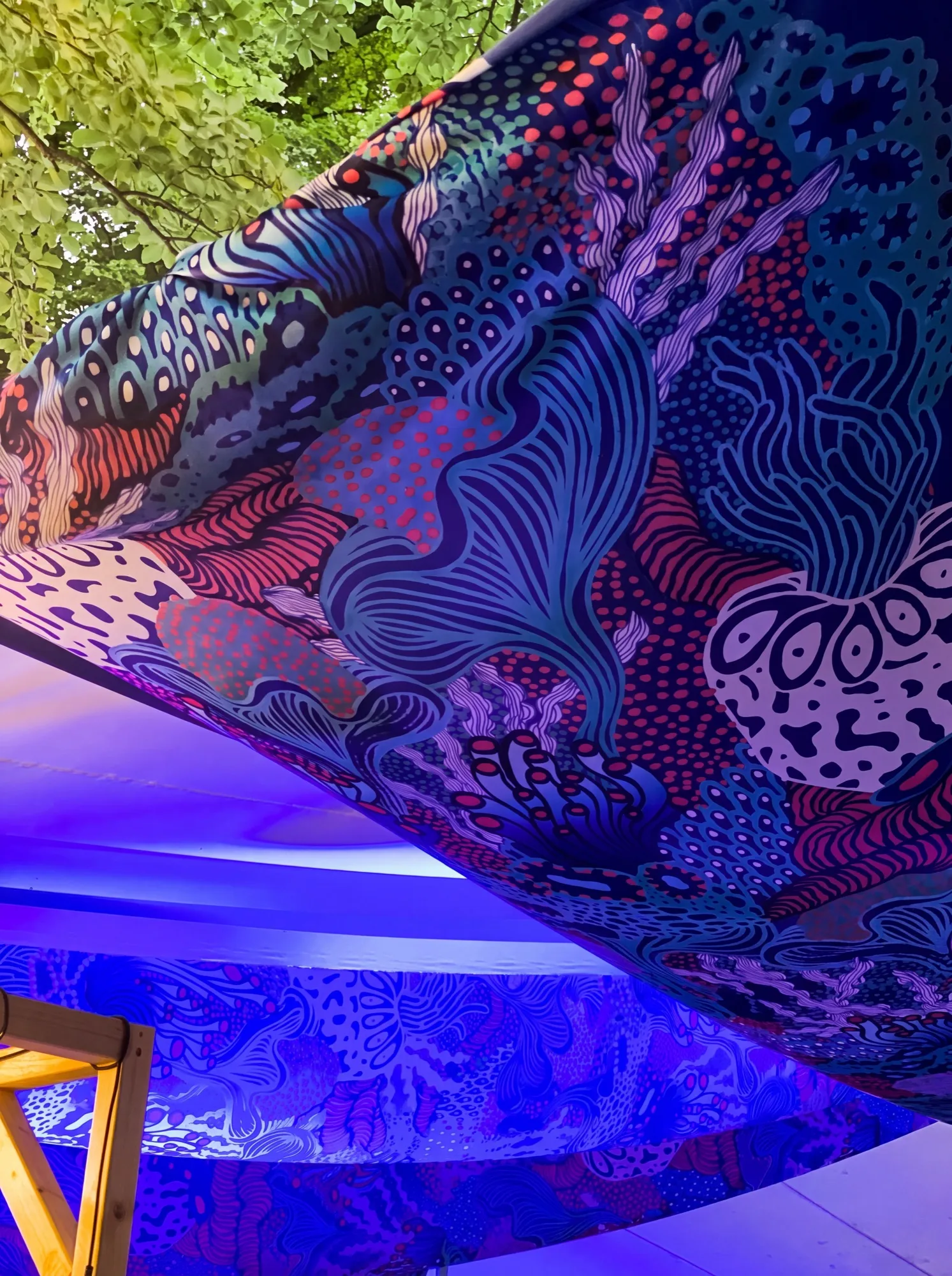 Close-up of Coral Garden forming a fabric canopy beneath green foliage, with seaweed-like forms, rings and dotted textures in blue, coral and pale purple.