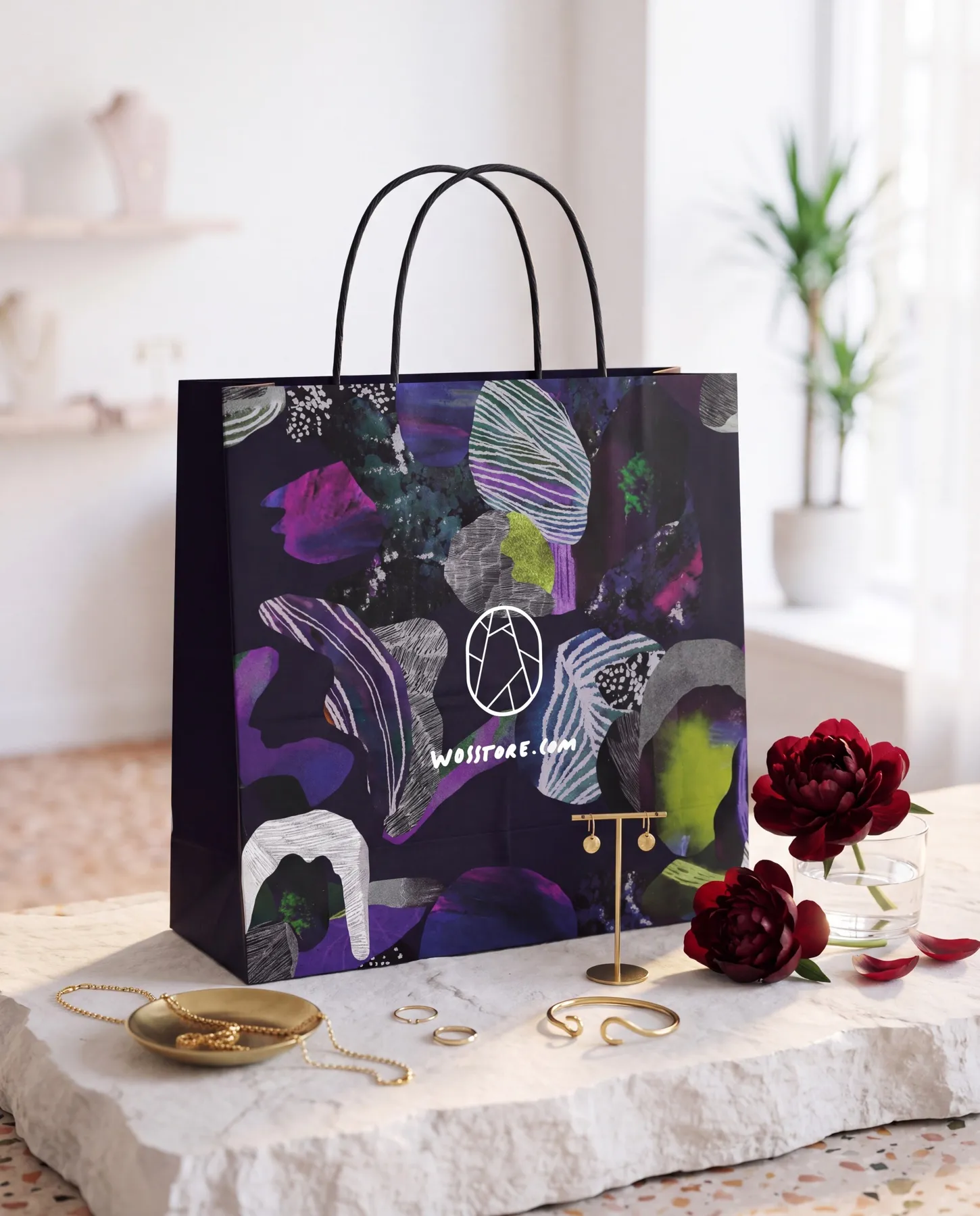 Mörkblå papperspåse med svarta tvinnade handtag på en ljus stenskiva bland guldsmycken och vinröda pioner: påsen bär abstrakta mineral- och stenformer i lila, magenta, limegrönt och grått med vita streckade strukturer, och i vitt WOS-symbolen — en tecknad ädelsten — över texten wosstore.com — uppdrag av Amandah L'Narte för WOS