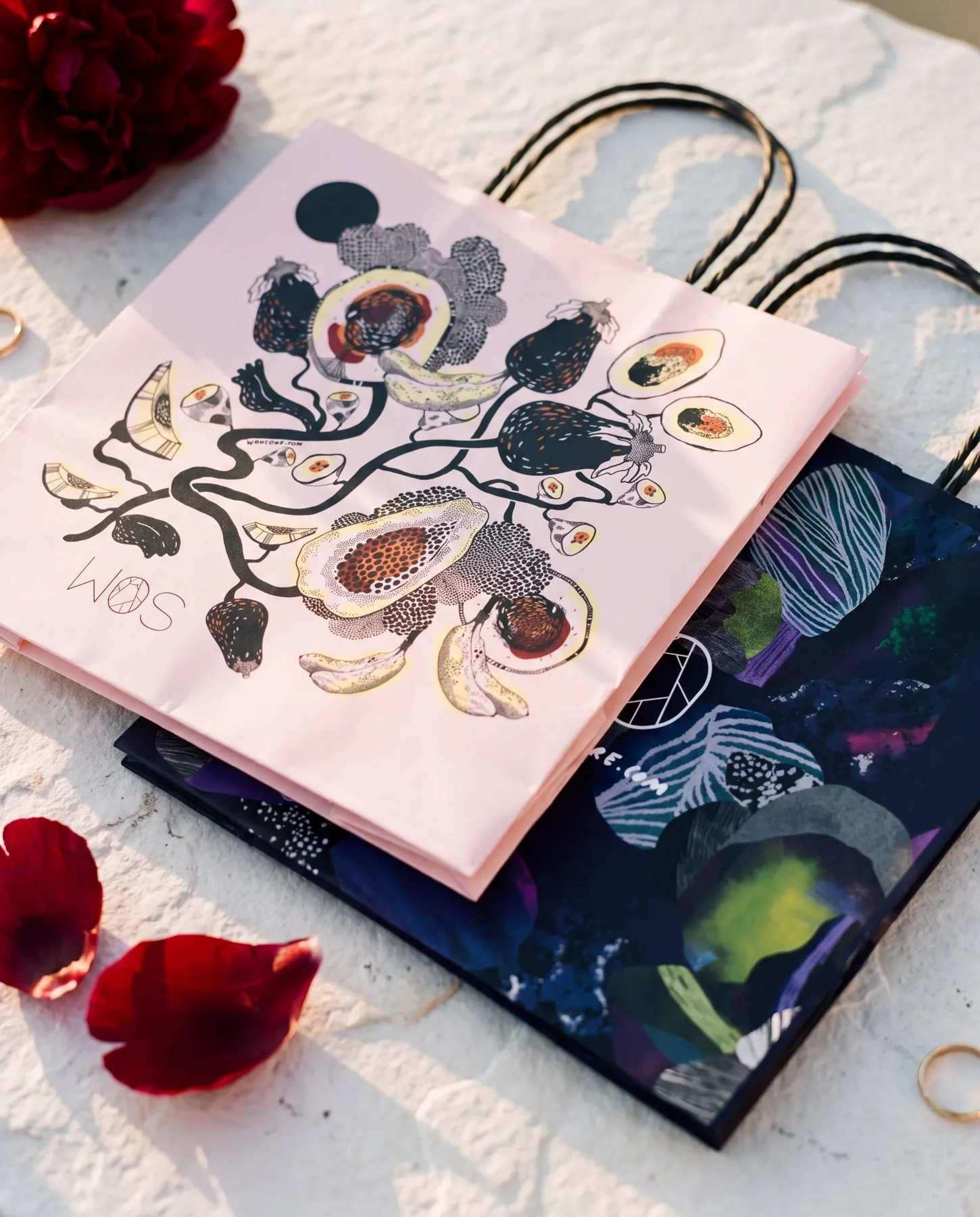 Båda butikspåsarna liggande omlott på en vit stenyta med vinröda pionblad omkring — den ljusrosa med fruktillustrationen överst och den mörkblå med mineralformerna under — uppdrag av Amandah L'Narte för WOS