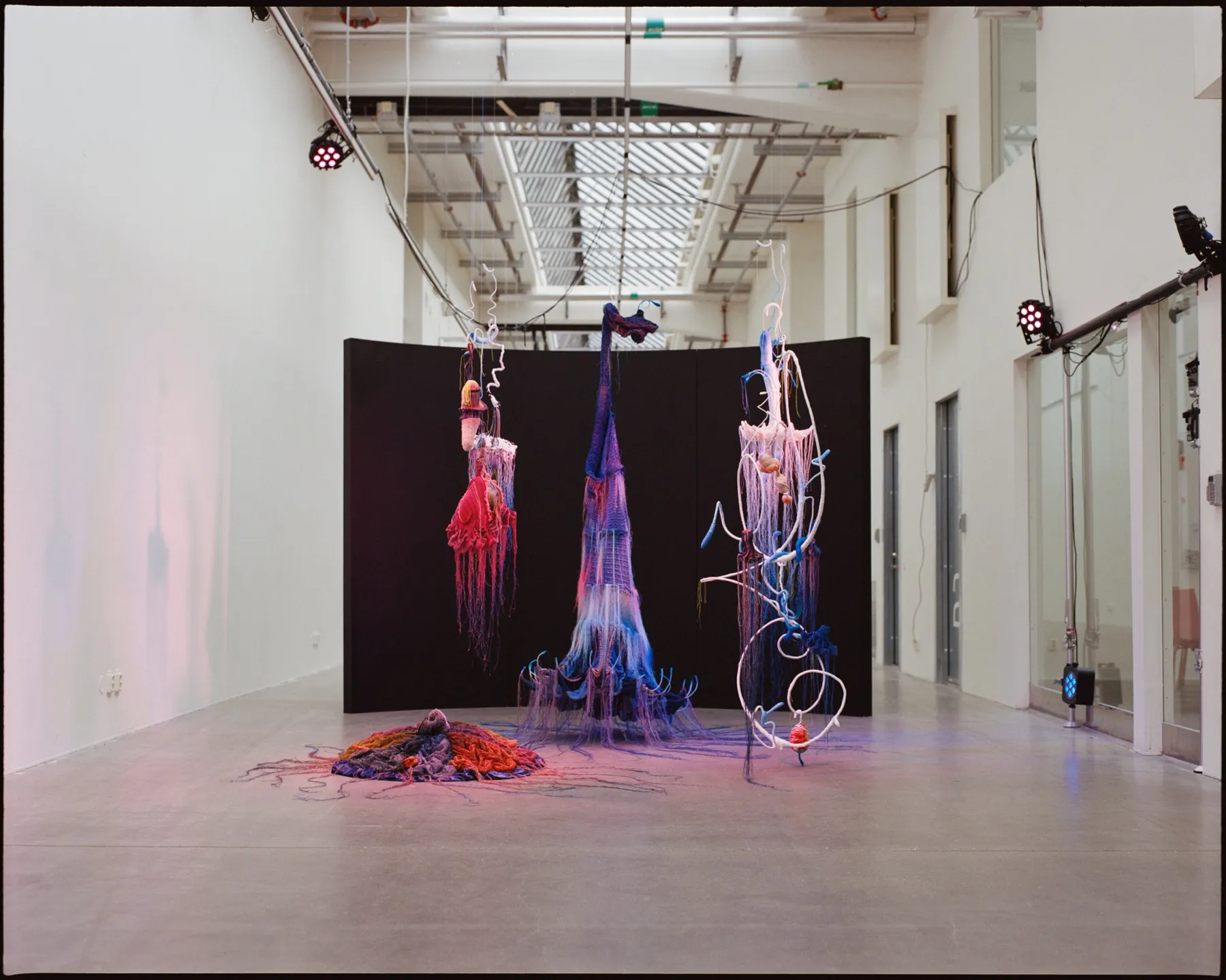 Installationsvy från Galleri Konstfack 2022 — tre hängande textilskulpturer i lila, blått och rosa med långa fransar svävar framför en svängd svart fondvägg i det vita gallerirummet, och en rund textil i rött, orange och lila ligger utbredd på golvet