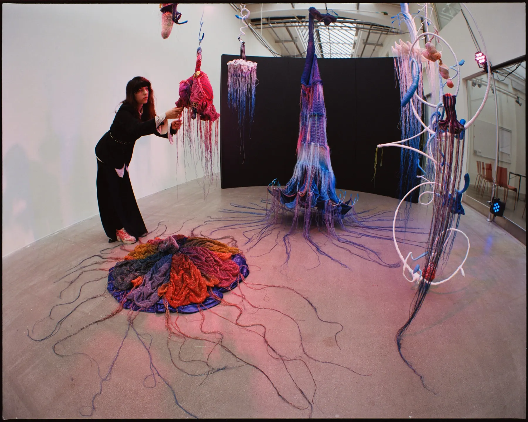 En person i svarta kläder justerar en hängande röd-rosa textilform i installationen — runt personen den fransade skulpturen i lila och blått, en svart fondvägg och en rund golvtextil i rött, orange och lila med utlöpande trådar