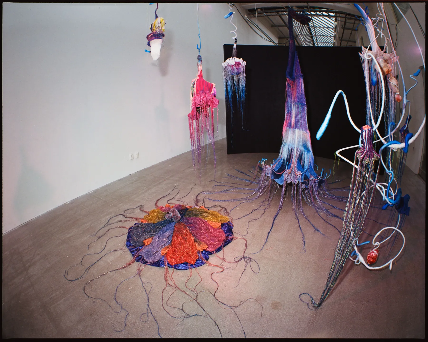 Installationsvy från sidan — hängande textilskulpturer i rosa, lila och blått med långa fransar framför en svart fondvägg, slingrande vita och blå vadderade linor till höger och den runda golvtextilen i rött, orange och lila i förgrunden