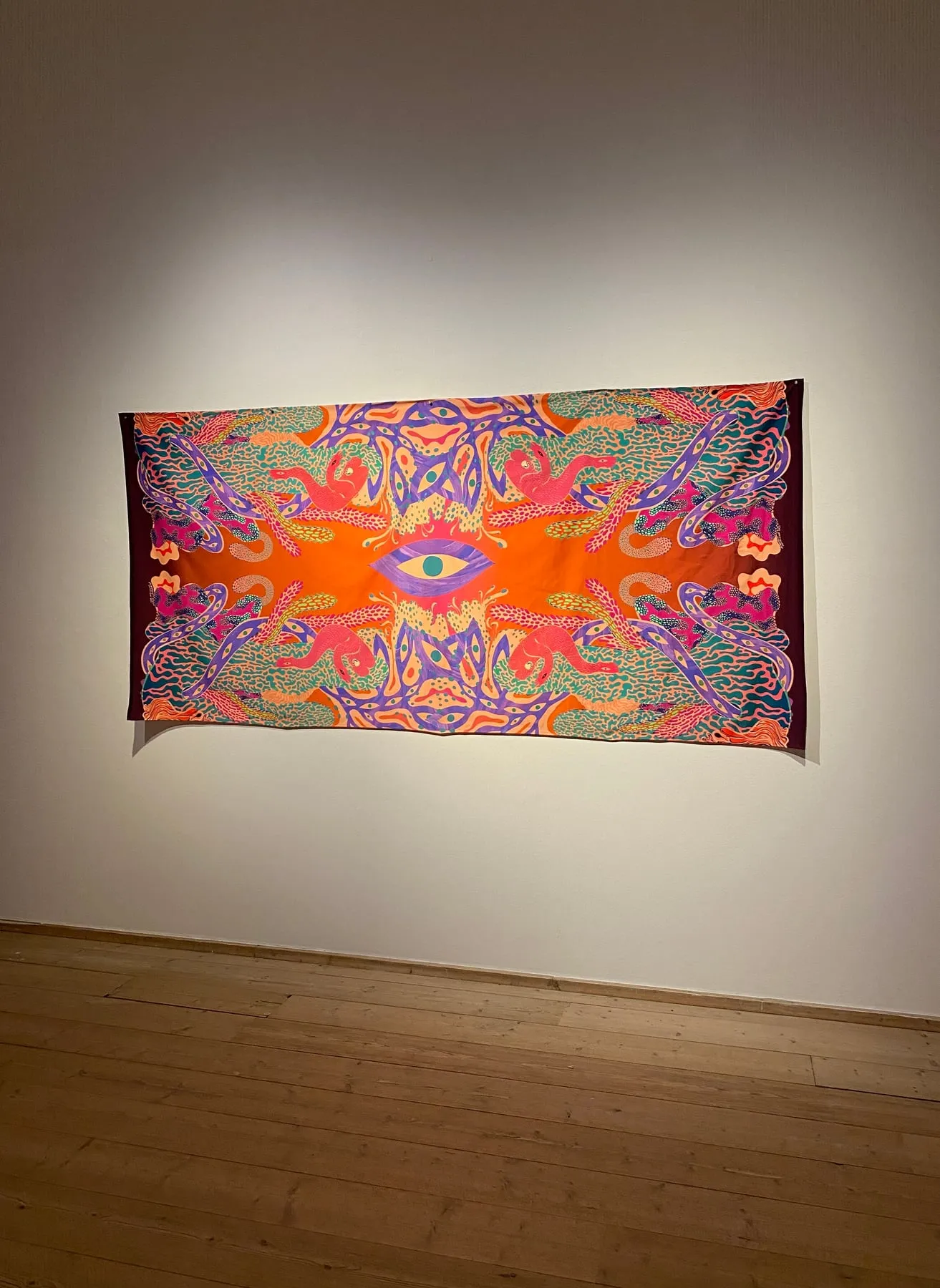 Tryckt textil på vit vägg i Millesgårdens konsthall — spegelvänt motiv i orange, lila och rosa med ett stort öga i mitten, omgivet av röda ormlika varelser, korallformer och slingrande lila band med ögon — foto: Amandah L'Narte