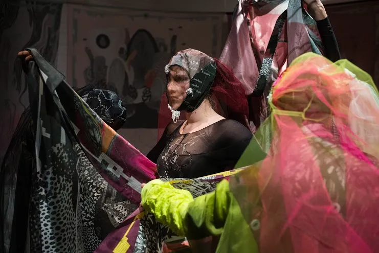Ur utställningen på Steneby Konsthall 2016 — en gestalt i dunkelt ljus, draperad i tyll och tryckta textilier med text i sjok av rosa, grönt och svart, framför ett mönstrat fondmotiv