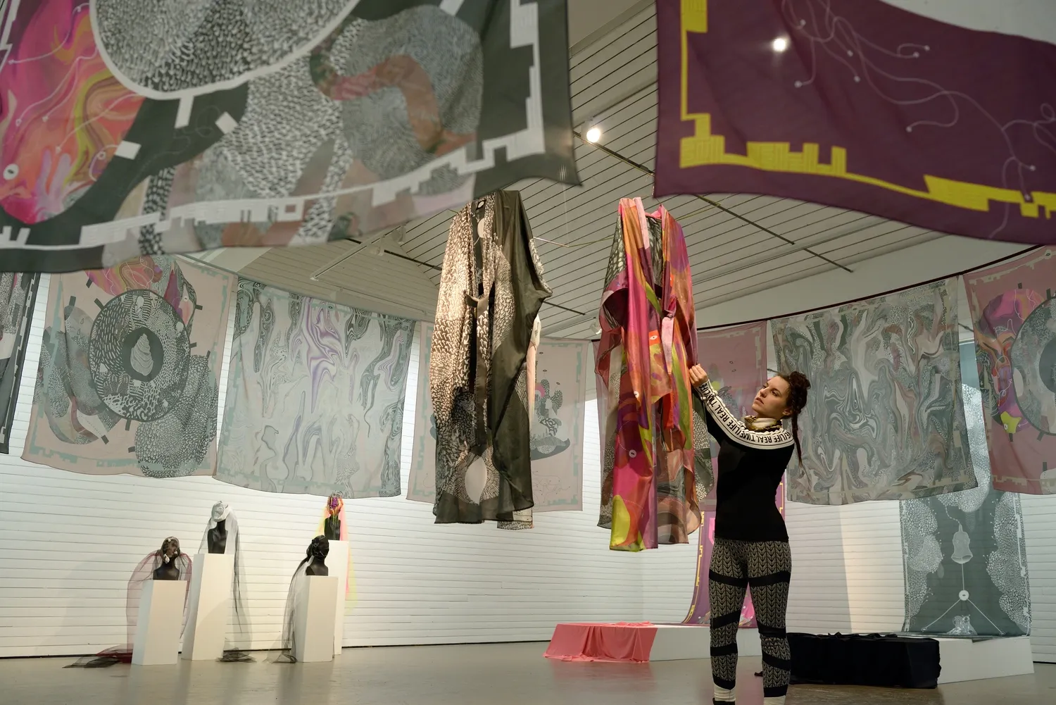 Installationsvy från Steneby Konsthall 2016 — tryckta textilier och sjalar hänger fritt i det vita rummet, plagg svävar från taket och Amandah L'Narte sträcker sig upp och justerar ett av dem; på vita podier i förgrunden står byster draperade i flor
