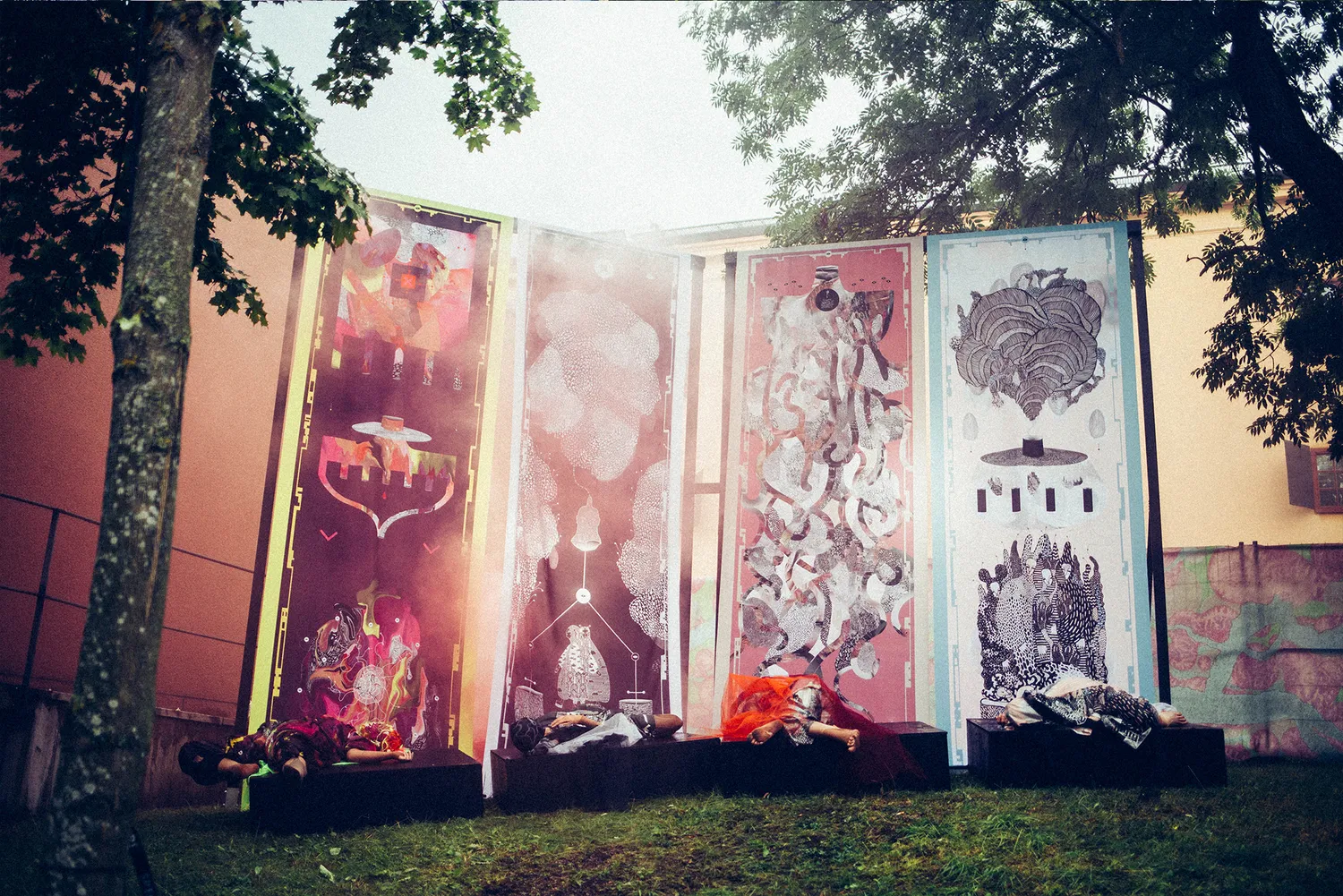 Fyra höga tryckta fanor står resta intill varandra i en park vid Stockholm Music & Arts 2016 — mönster i neongult, rosa, vinrött och svartvitt, med personer liggande på svarta podier framför, i rökigt motljus under träden