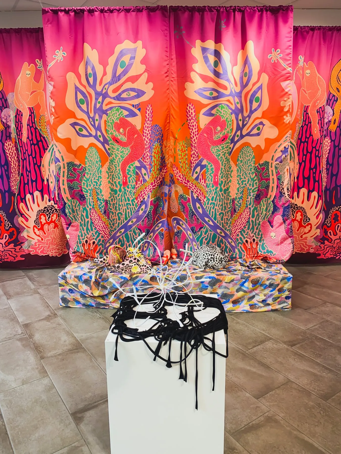 Installationsvy rakt framifrån — stora draperier i satin med spegelvänt motiv i rosa, orange och lila: korallika kaktusformer, röda ormlika varelser och persikofärgade väsen. Framför dem en låg plattform klädd i flerfärgat prickmönstrat tyg med leopardprickiga kuddar, och närmast en vit piedestal med ett nystan av svart snodd och en vit trådskulptur — foto: Amandah L'Narte