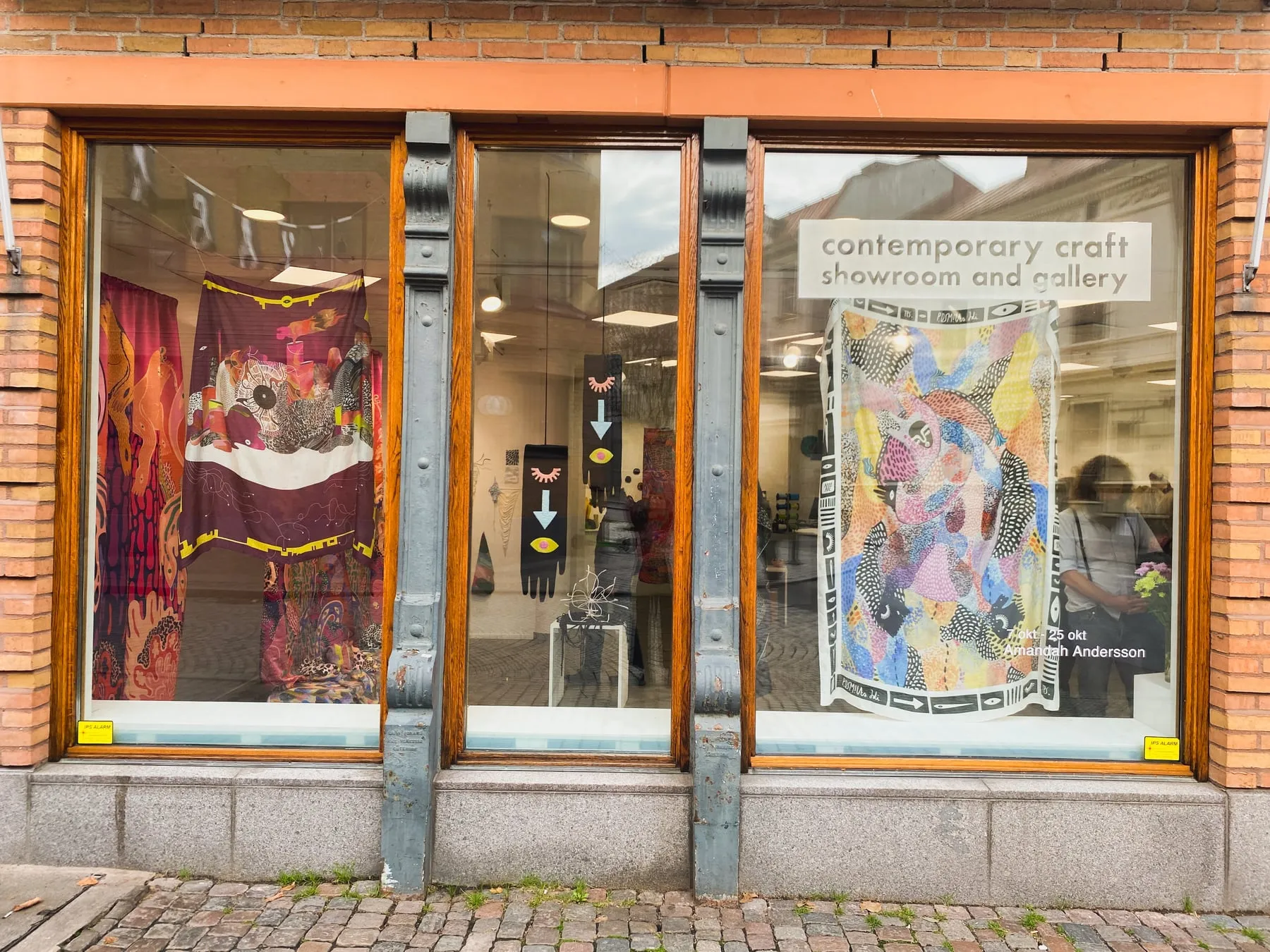 Galleri Sintras skyltfönster från gatan — i fönstren hänger ett vinrött tyg med gul bård och ett flerfärgat lapptäckslikt tyg, med skylten contemporary craft showroom and gallery och texten 7 okt–25 okt Amandah Andersson — foto: Amandah L'Narte