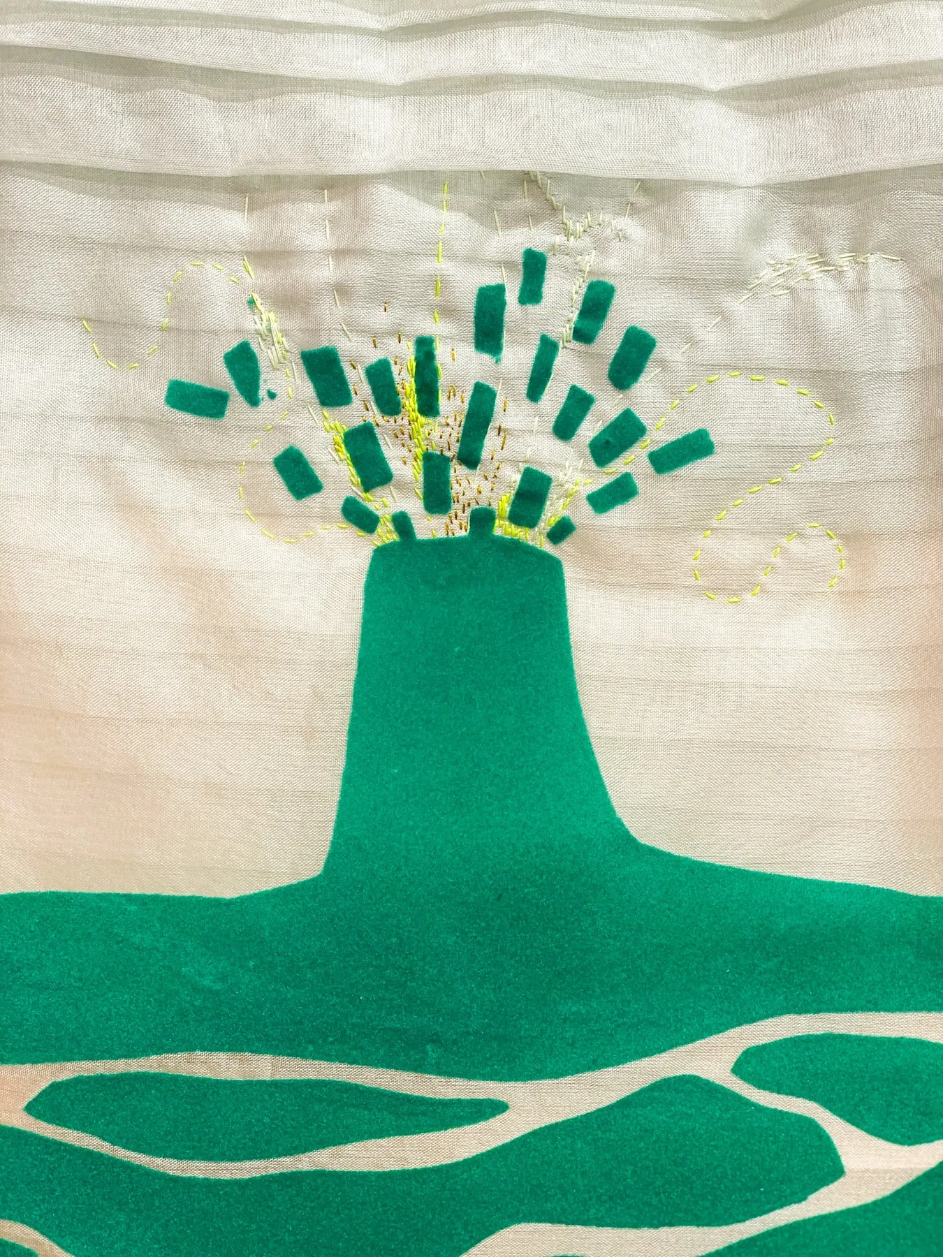 Detalj av ett litet textilt verk — en grön vulkanform i flock som sprutar ut gröna filtbitar, omgiven av broderade stygn i gult och guld på skirt veckat tyg i vitt och beige — foto: Amandah L'Narte